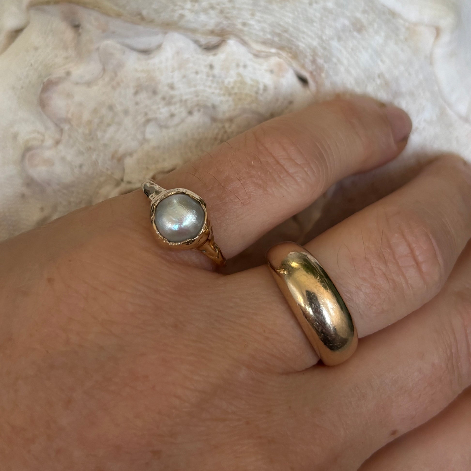 Close-up of a hand wearing two gold rings with a pearl on a textured surface.